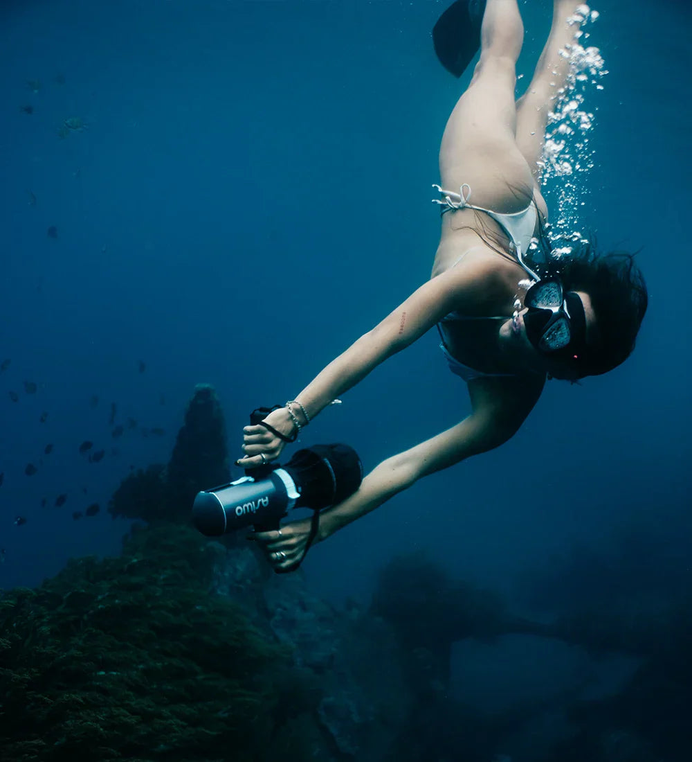 A freediver in red swim trunks propels through clear blue water using a black Asiwo U1 underwater scooter.