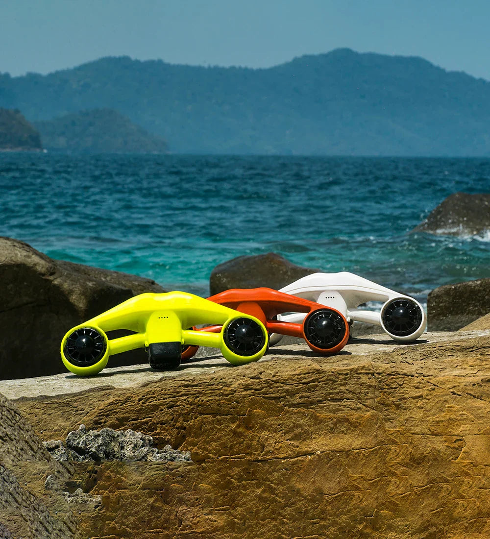 Three ASIWO MANTA underwater sea scooters in yellow, orange, and white, displayed on a rock near the ocean with mountains in the background