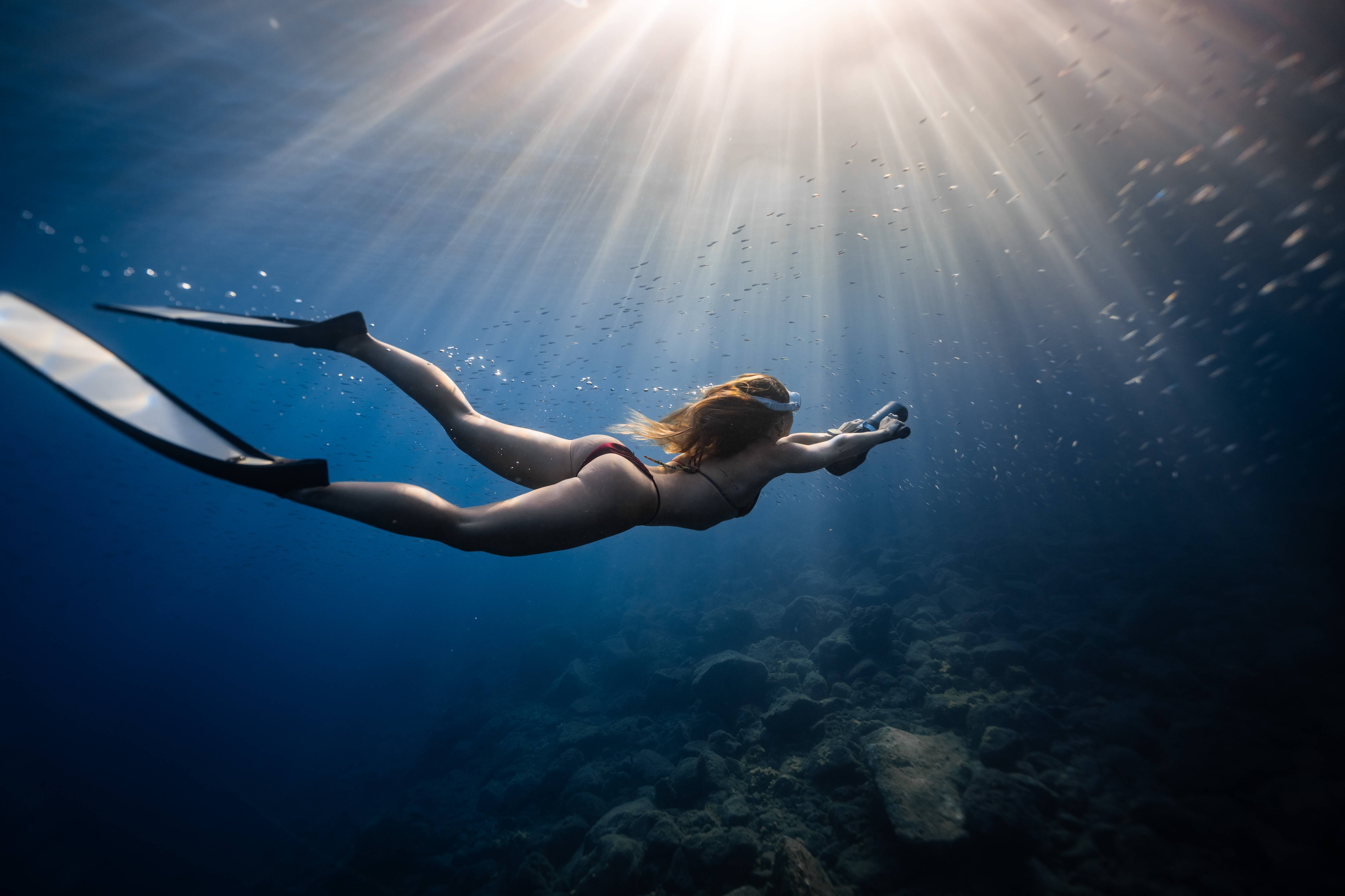 Snorkeler wearing long fins swimming underwater toward sunlight over a rocky seabed