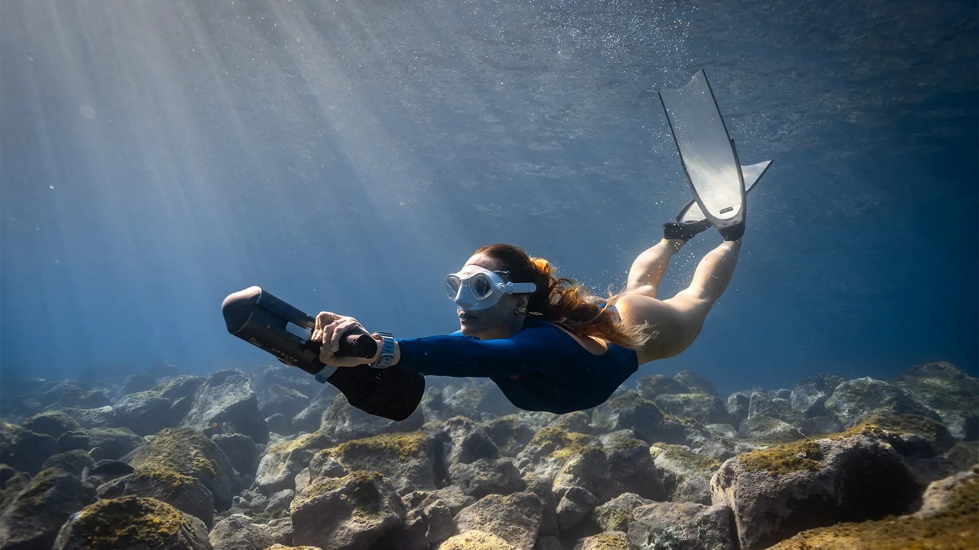 Woman scuba diving with the ASIWO U1 Diver Propulsion Vehicle over a rocky seabed in clear blue water.