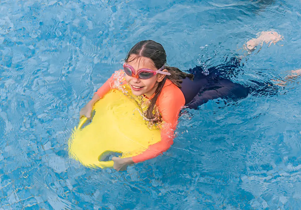 Child using an ASIWO Mako electric kickboard in a swimming pool, holding the yellow board while gliding on the water with goggles on