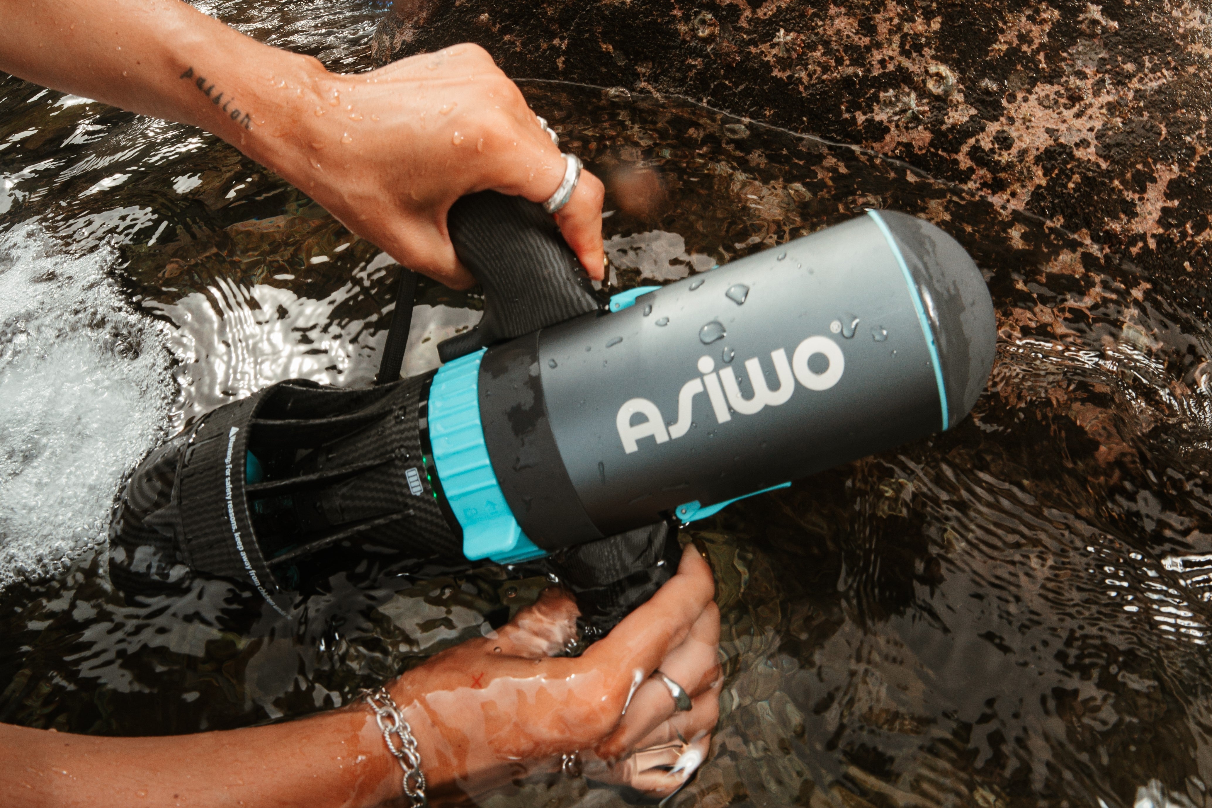 Close-up of hands holding an ASIWO diver propulsion vehicle in shallow seawater near coastal rocks.