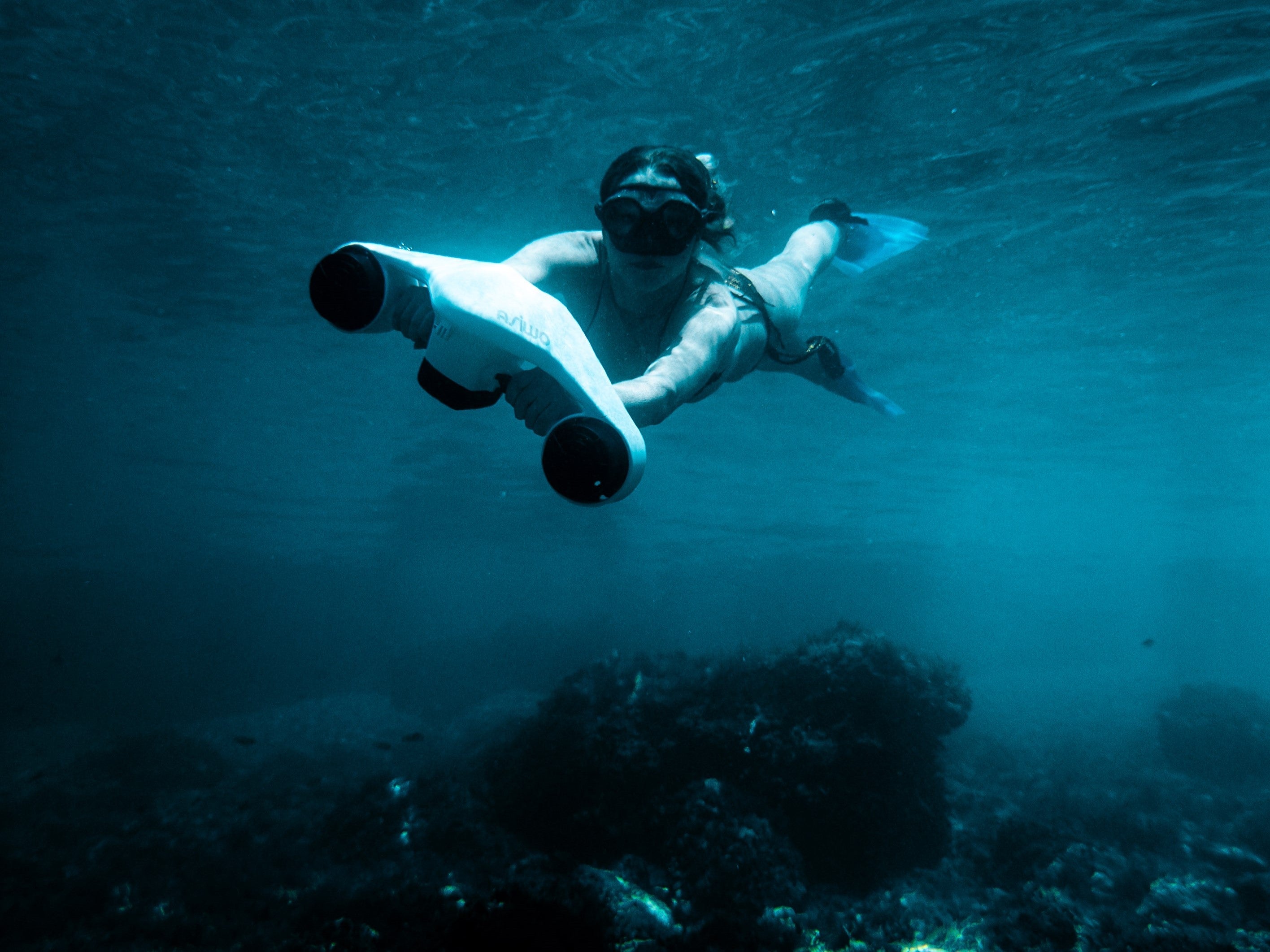 Diver swimming underwater with a white ASIWO Manta underwater scooter above a reef in clear blue ocean water.
