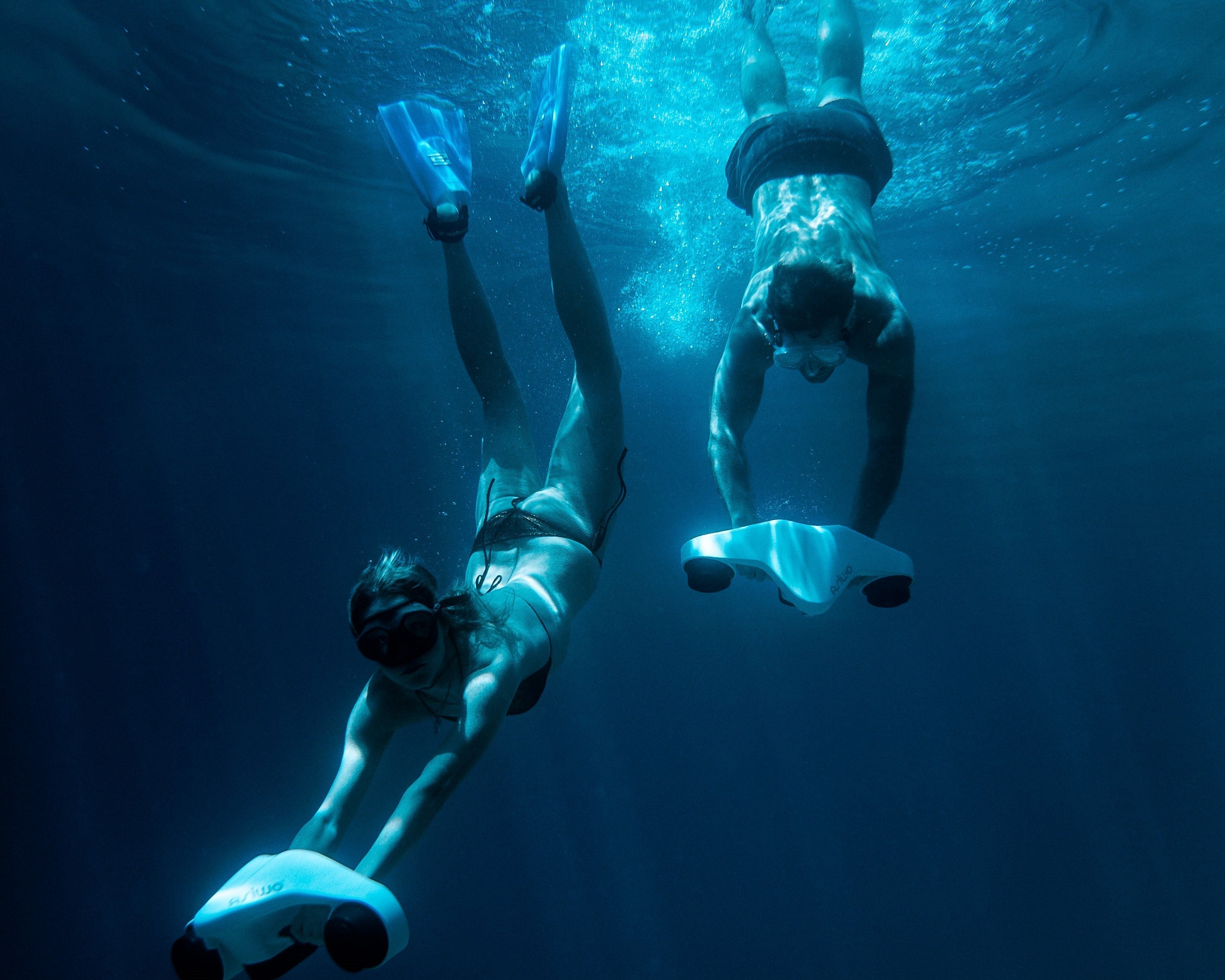 Two swimmers underwater using ASIWO Manta underwater scooters, diving together in clear blue ocean water