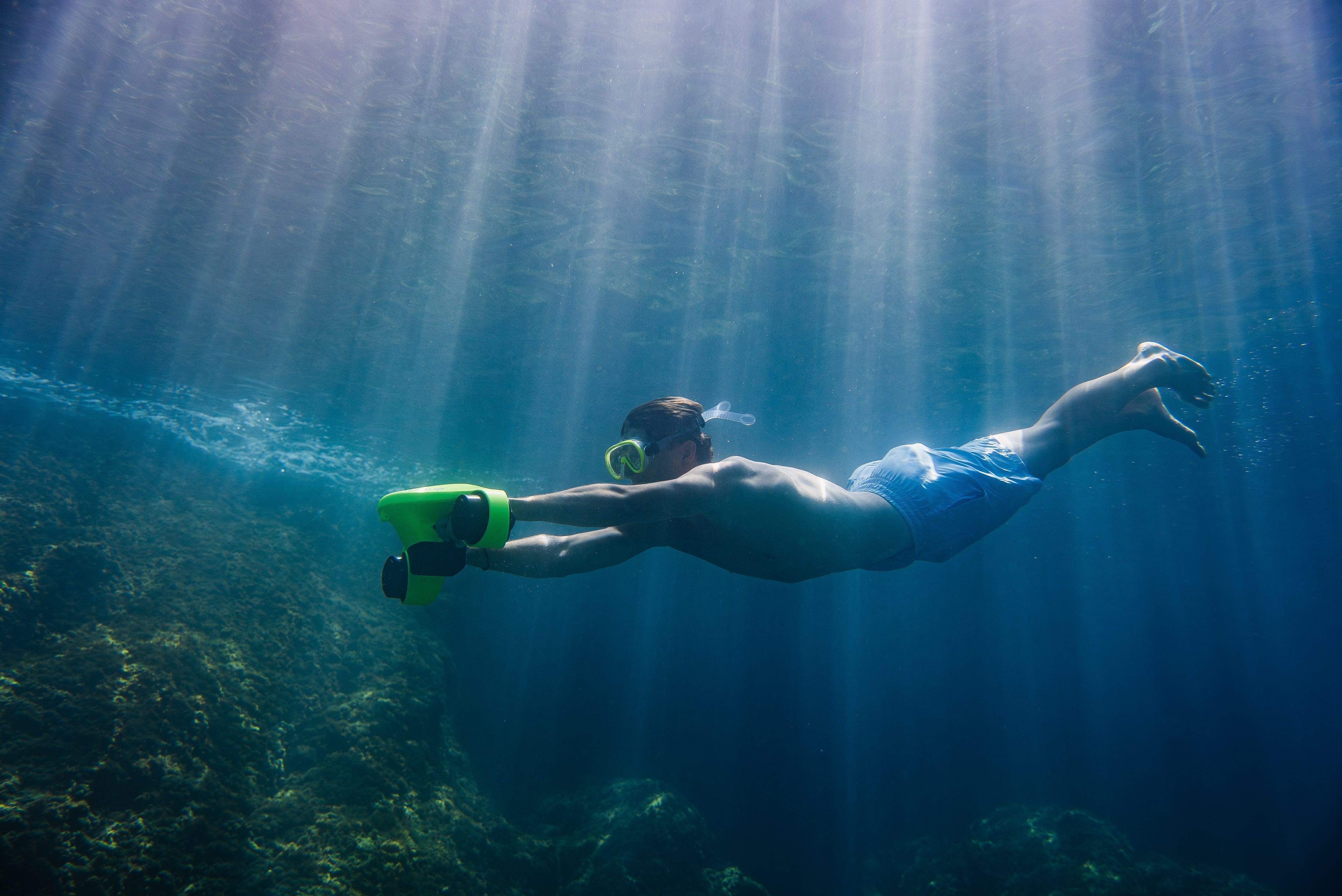 Snorkeler gliding underwater with an ASIWO Manta underwater scooter, swimming above a rocky reef as sun rays stream through clear blue water.