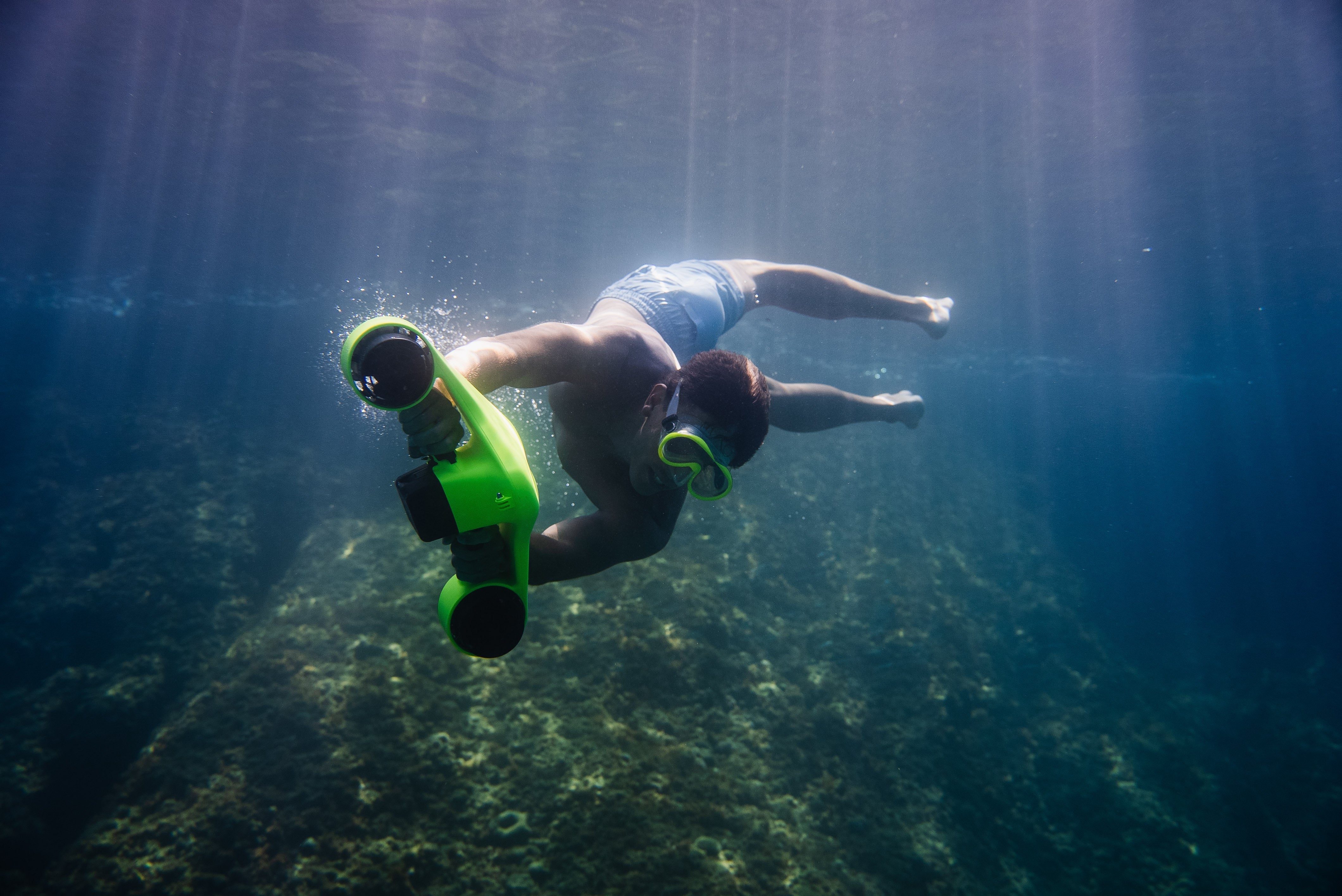 Snorkeler exploring a reef underwater with a green ASIWO Manta underwater scooter, swimming above the ocean floor in clear blue water.