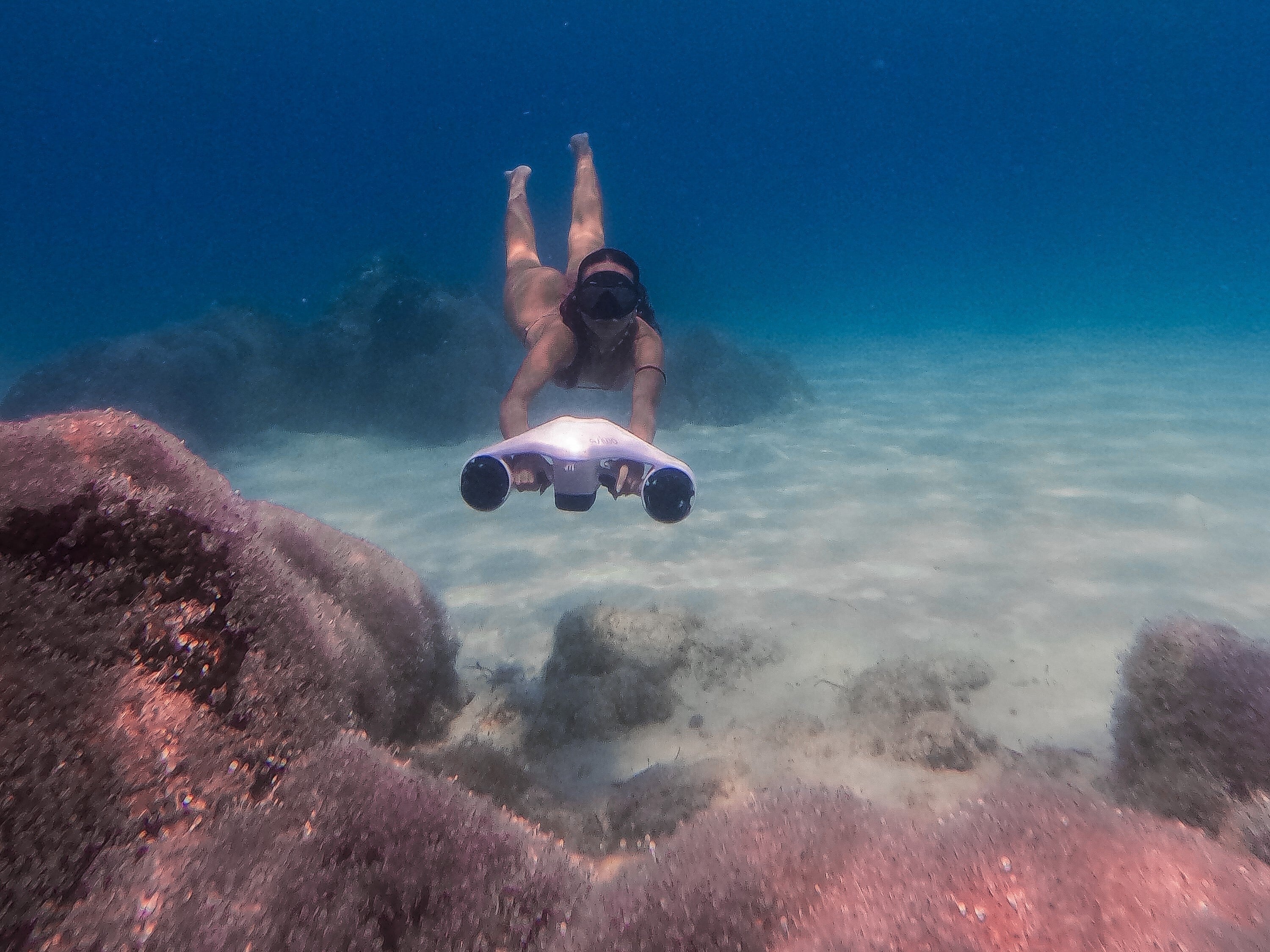 Snorkeler using an underwater scooter to swim over a sandy seabed with rocks in clear ocean water