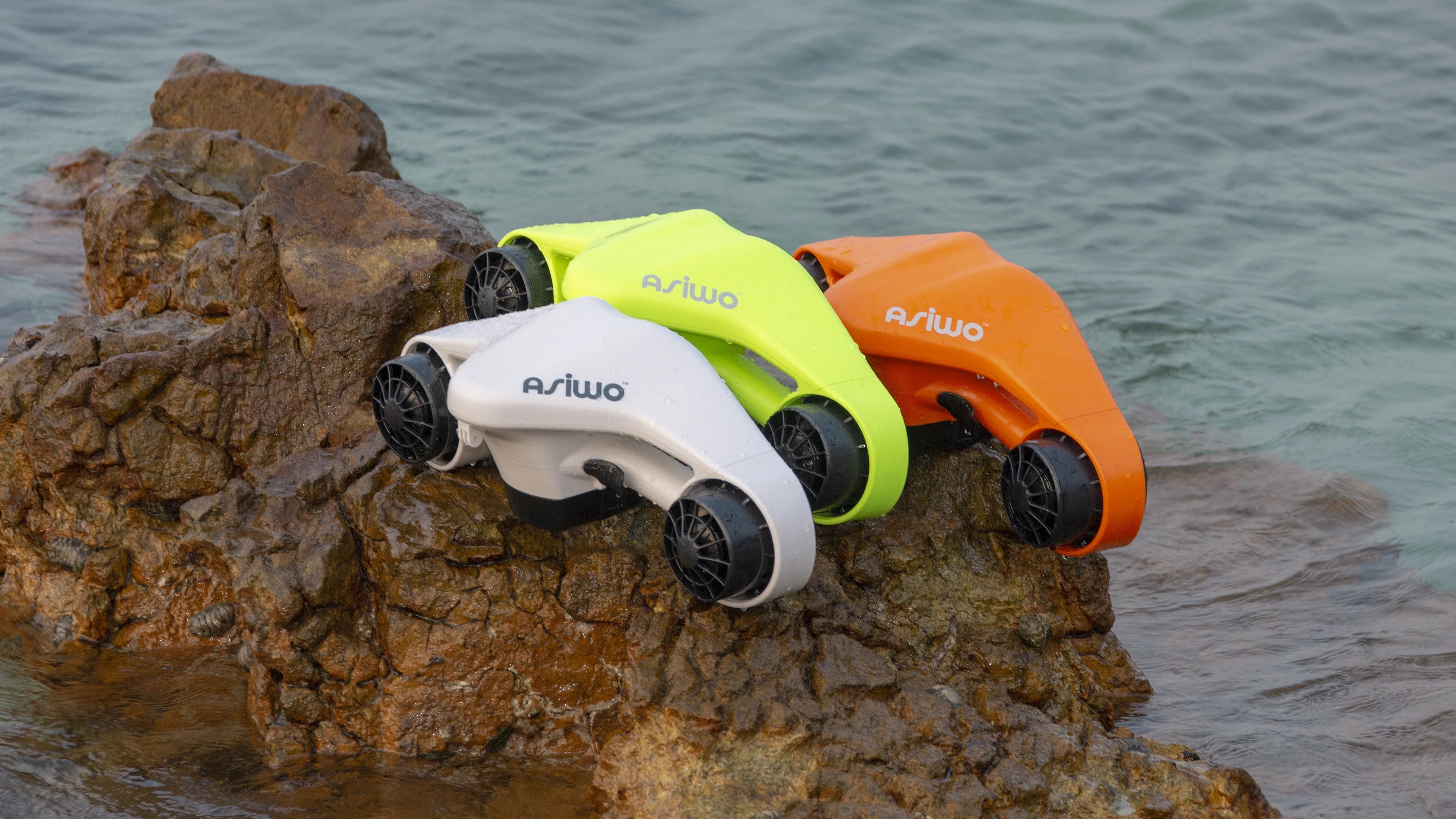 Three underwater scooters in white, green, and orange resting on a rock by the sea.