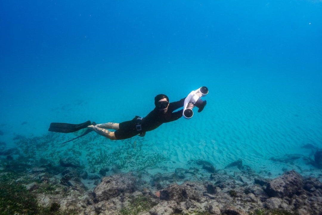 ASIWO Manta underwater scooter propelling a diver over a clear sandy seabed with rocks and blue ocean water