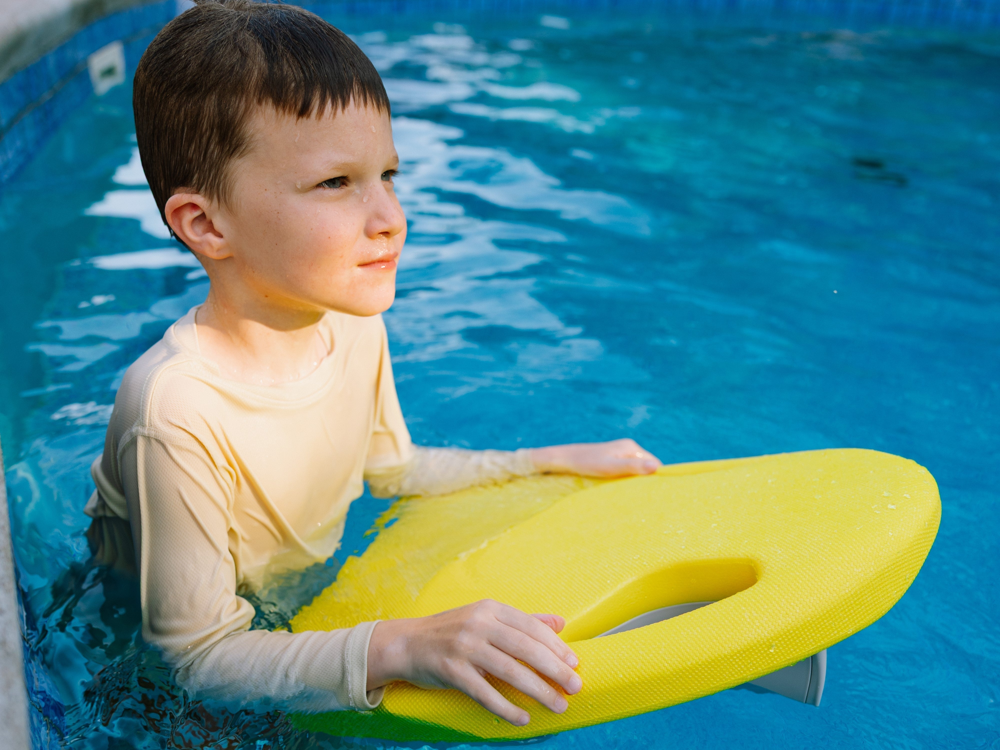 A child standing in a swimming pool holding a yellow electric kickboard.